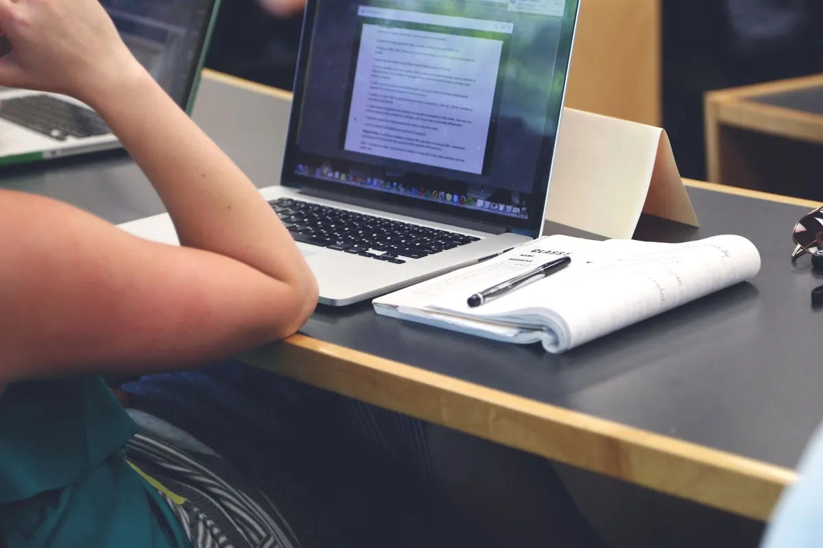 A student in a classroom using a laptop while writing notes in a notebook.